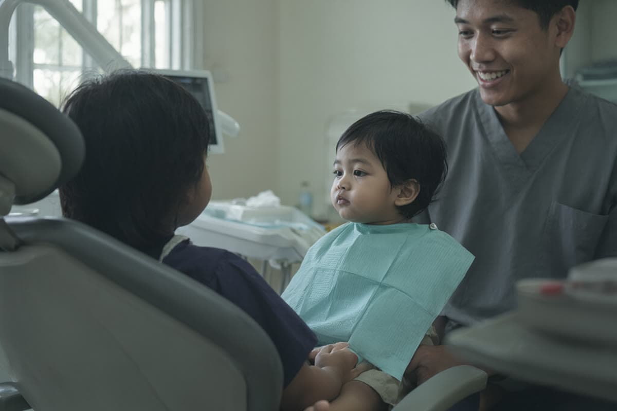 A young Malaysian child sitting on a parent's lap in a dental chair while a dentist smiles