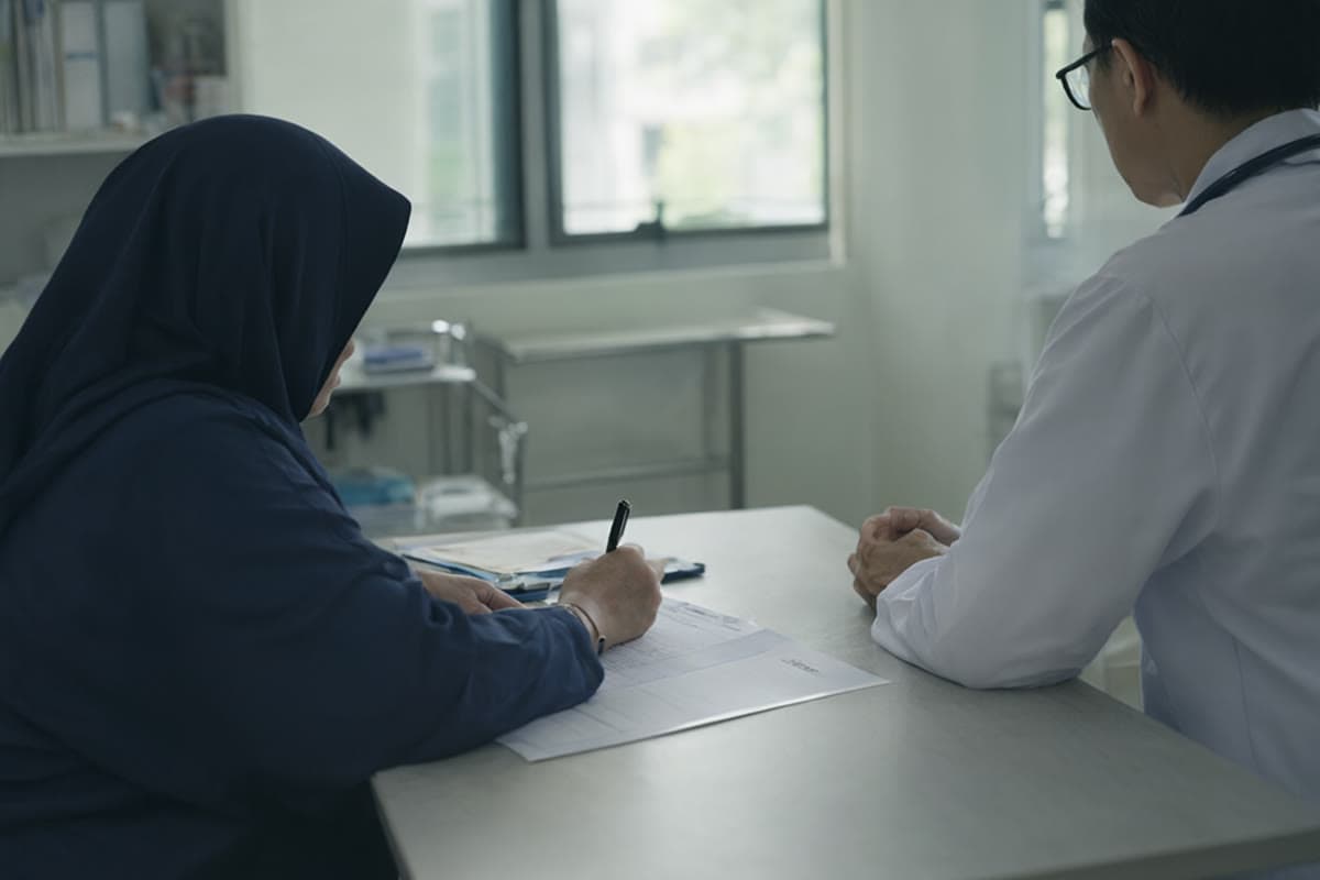 A patient writing a food and symptom diary at a clinic table with a doctor