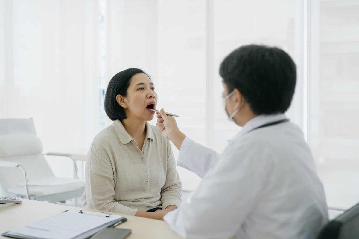 A patient seated in a bright clinic consultation room while a doctor inspects the throat